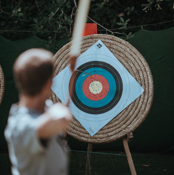 Eine Person zielt mit einem Bogen auf eine aufgestellte Zielschreibe, in der Zielscheibe steckt ein Pfeil.
