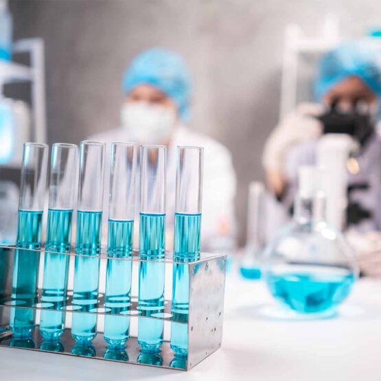 Reagenzgläser gefüllt mit blauer Flüssigkeit stehen auf einem Tisch in einem Labor, dahinter sind unscharf Laboranten an einem 