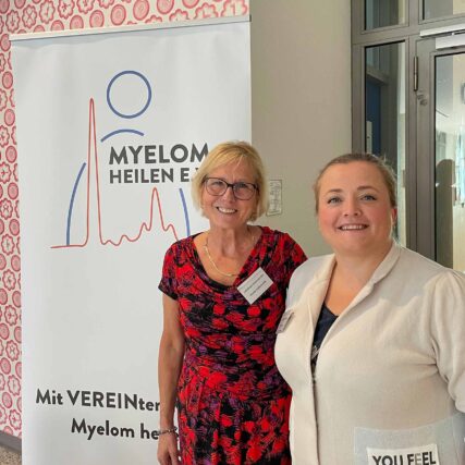 Zwei weibliche Vereeinsmitglieder stehen vor einem ca. 1,80m hohen Plakat des Vereins "Myelom Heilen e. V." mit dem Logo des Vereins und der Aufschrift "Mit VEREINten Kräften Myelom heilen!"