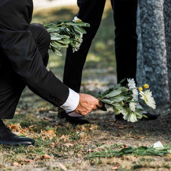 Ein Mensch in Trauerkleidung legt Blumen auf einen Waldboden.
